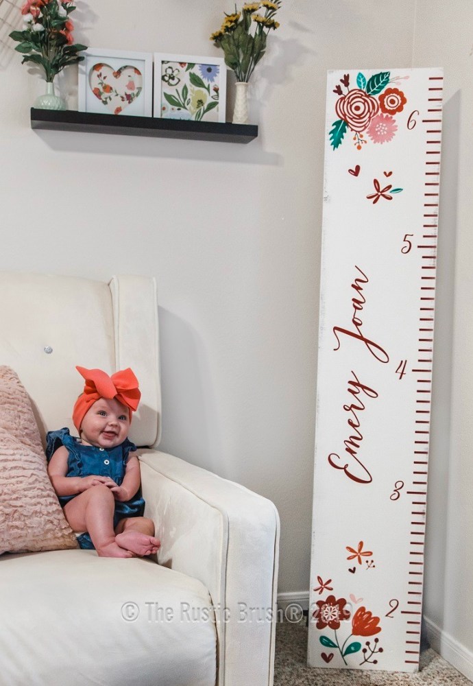 Baby in armchair beside floral growth chart, decorated shelf above.