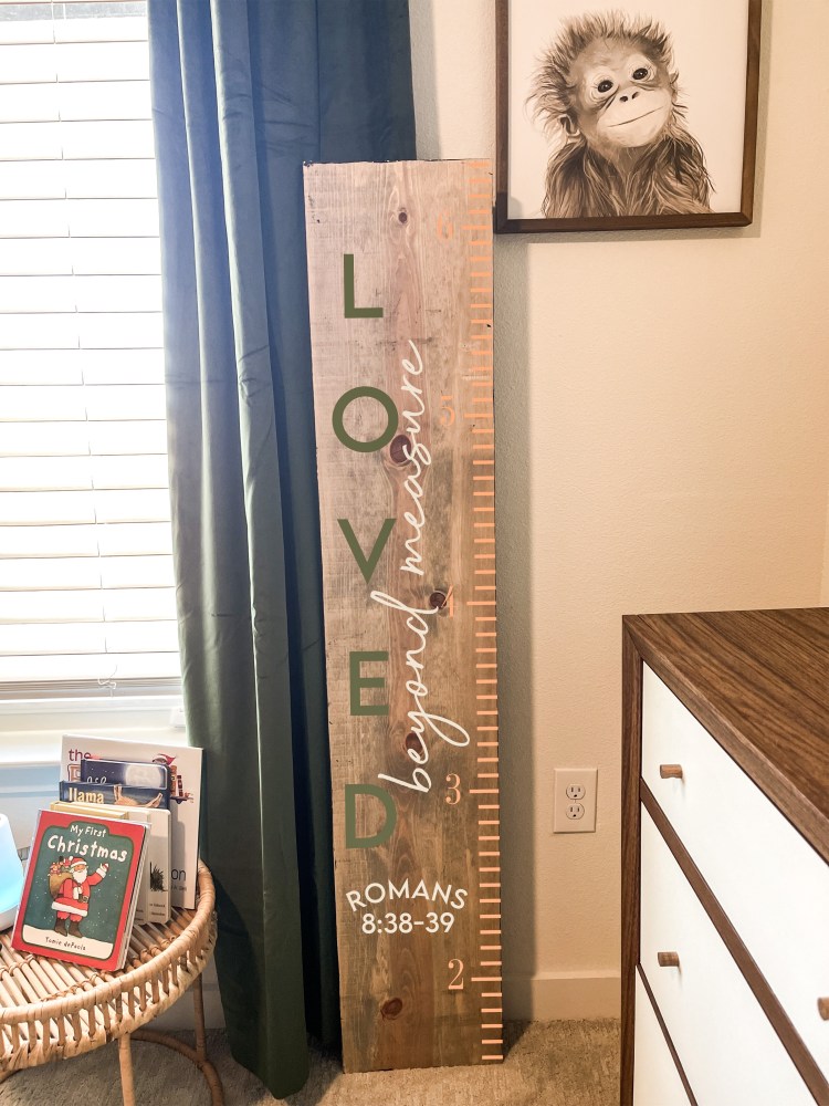 Wooden growth chart with 'Loved beyond measure' text, framed monkey art, and books on a wicker table.