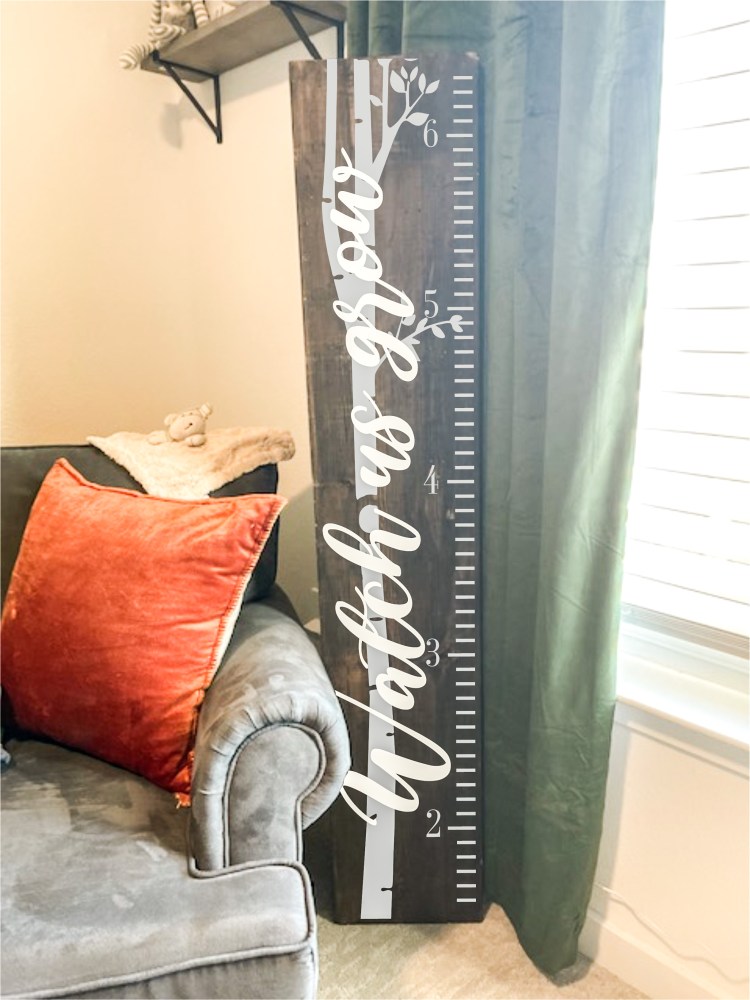 Wooden growth chart in room with 'Watch us grow' text, next to gray couch with orange pillow.