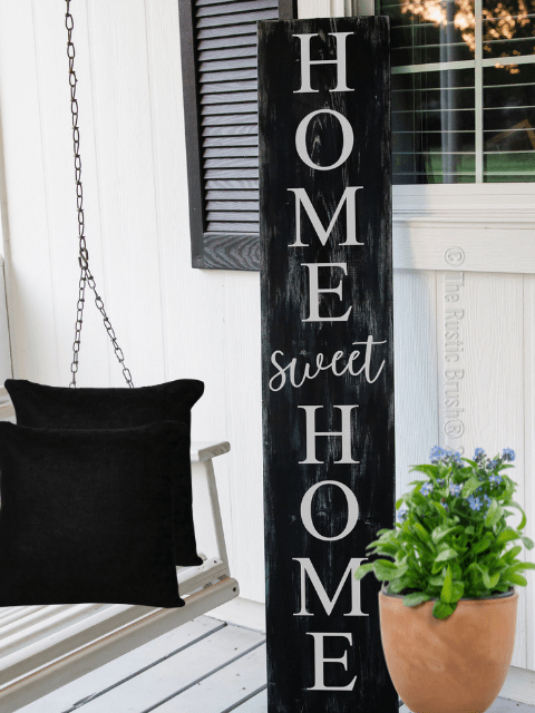 Porch with 'Home Sweet Home' sign, black pillows on a swing, and potted plant.