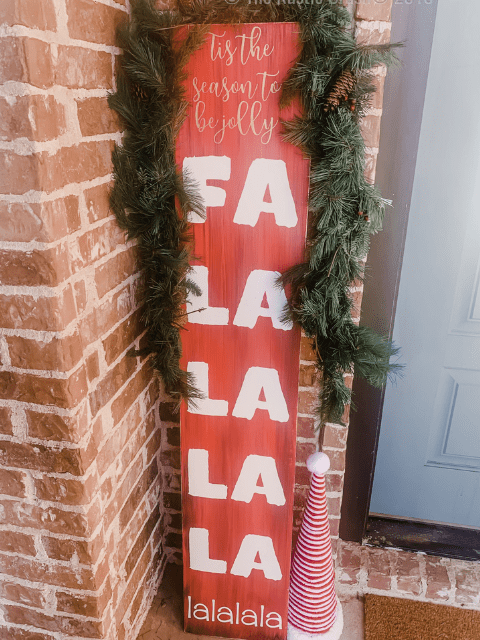 Red holiday sign with 'Tis the season to be jolly' and 'Fa la la la la' next to a brick wall and door.