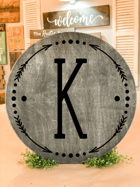 Wooden round sign with the letter 'K' in the center, surrounded by decorative border, on a stand.