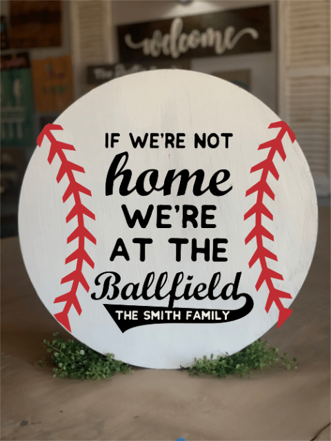 Round sign with baseball stitching, text says 'If we're not home, we're at the ballfield, The Smith Family'.
