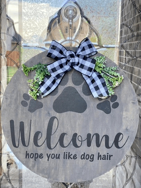 Round welcome sign with paw prints, bow, and greenery, saying: 'Welcome, hope you like dog hair.'