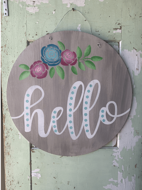 Round sign with 'hello' and painted flowers on rustic green and white background.
