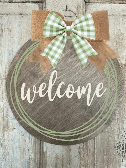 Wooden welcome sign with green bow and burlap accents on rustic door.