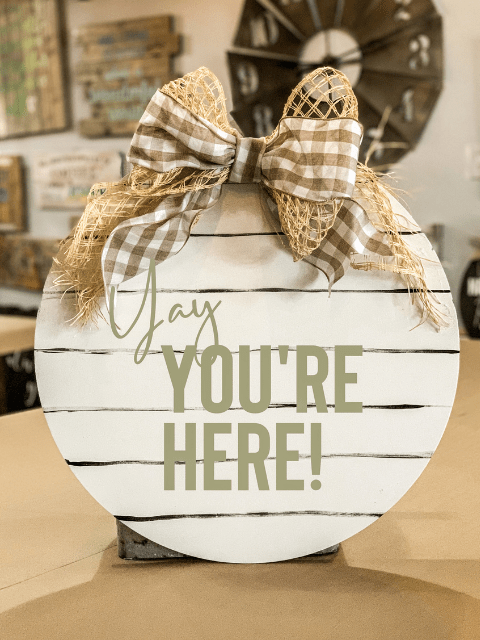 Round sign with checkered bow reads 'Yay YOU'RE HERE!' in a cozy indoor setting.