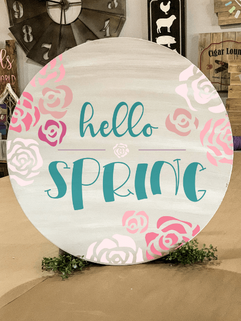 Round sign with 'hello spring' text and pink floral design on wood background.