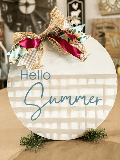 Round 'Hello Summer' sign with colorful bows and plaid background on a table.