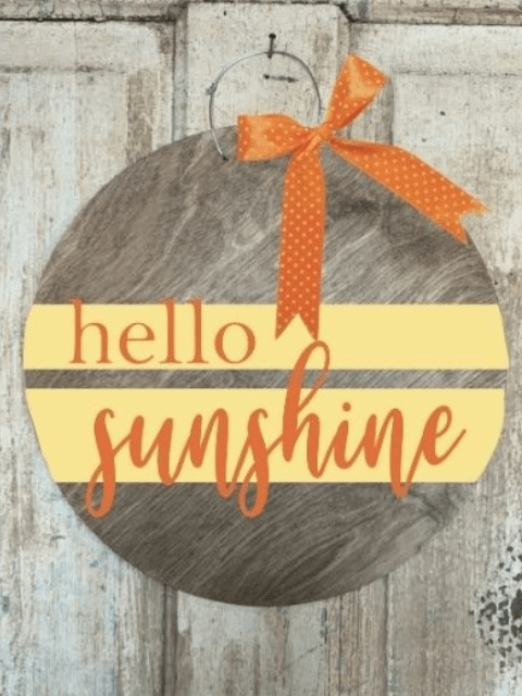 Round wooden sign with 'hello sunshine' and orange ribbon, hanging on a rustic background.