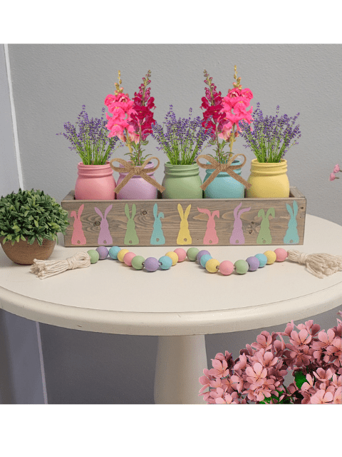 Pastel jars with flowers in a wooden box with bunny designs on a table with decorative beads.