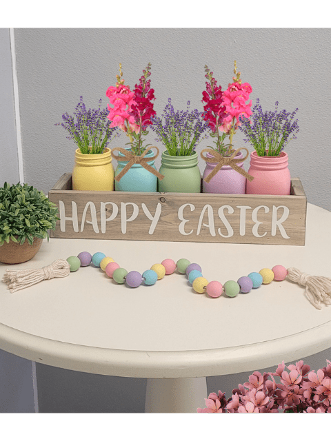 Colorful jars with flowers in a box labeled 'Happy Easter' on a table with pastel bead garland.
