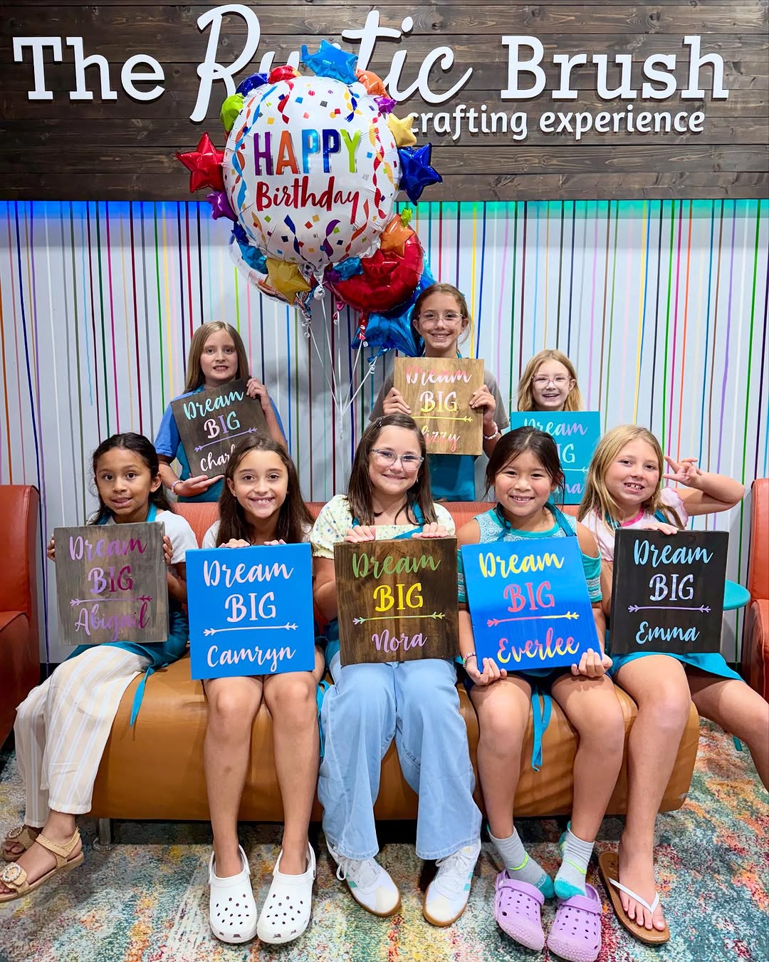 Eight girls holding 'Dream Big' signs with a birthday balloon at The Rustic Brush.