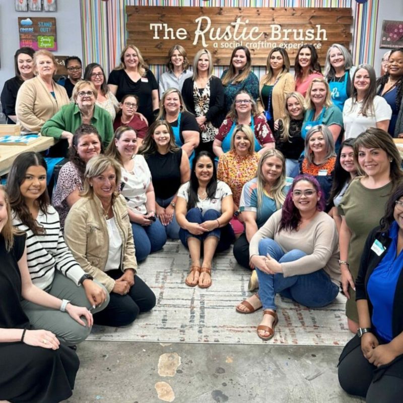 Three women painting a decorative board in a craft studio, wearing blue aprons labeled 'TRB'.