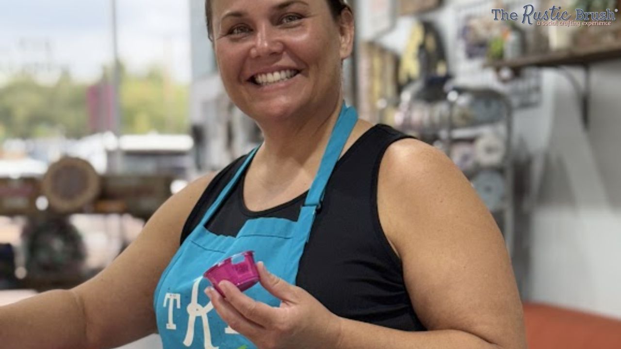 Smiling person wearing a blue apron holds a small object in a workshop setting.
