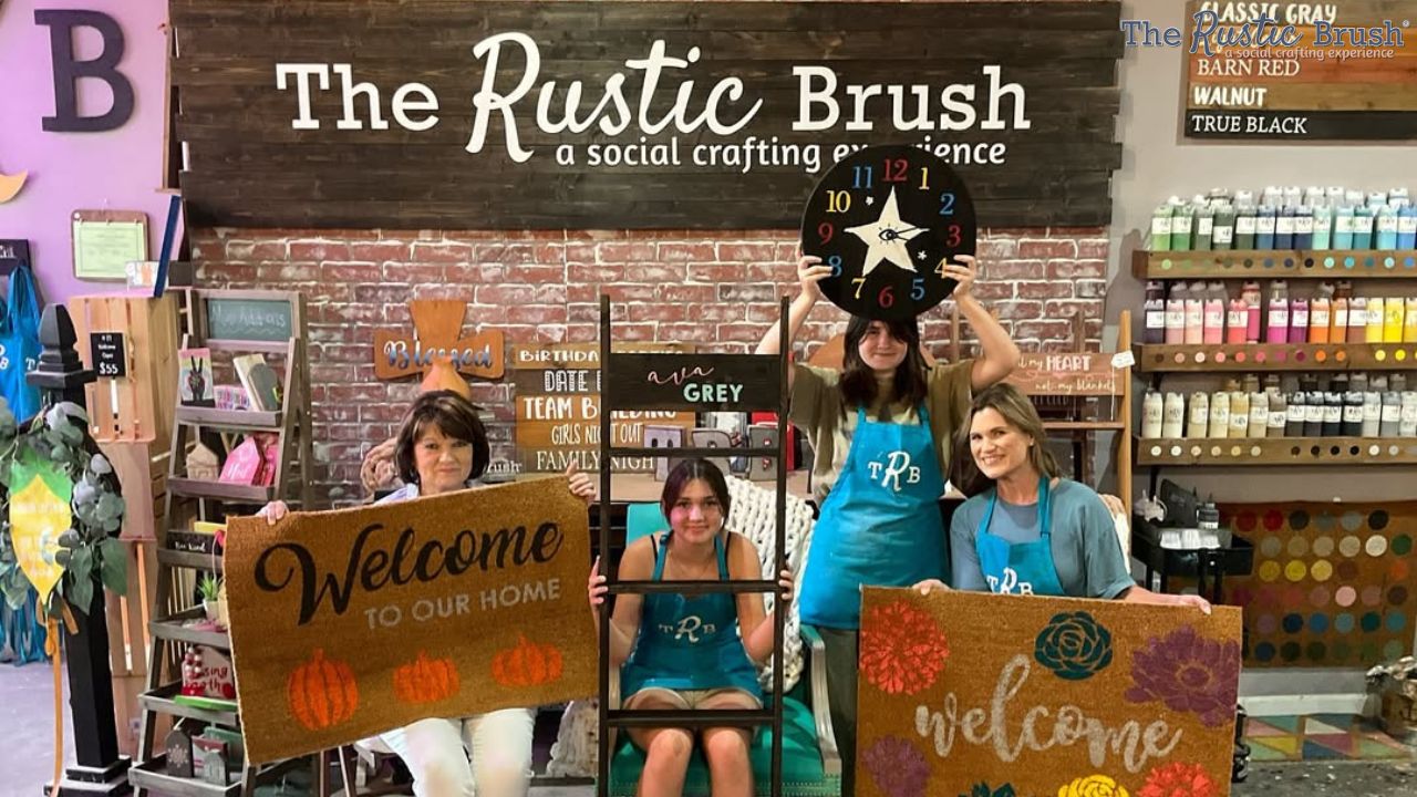 Four people at The Rustic Brush, holding crafts and signs, with a brick wall and paint shelves behind them.