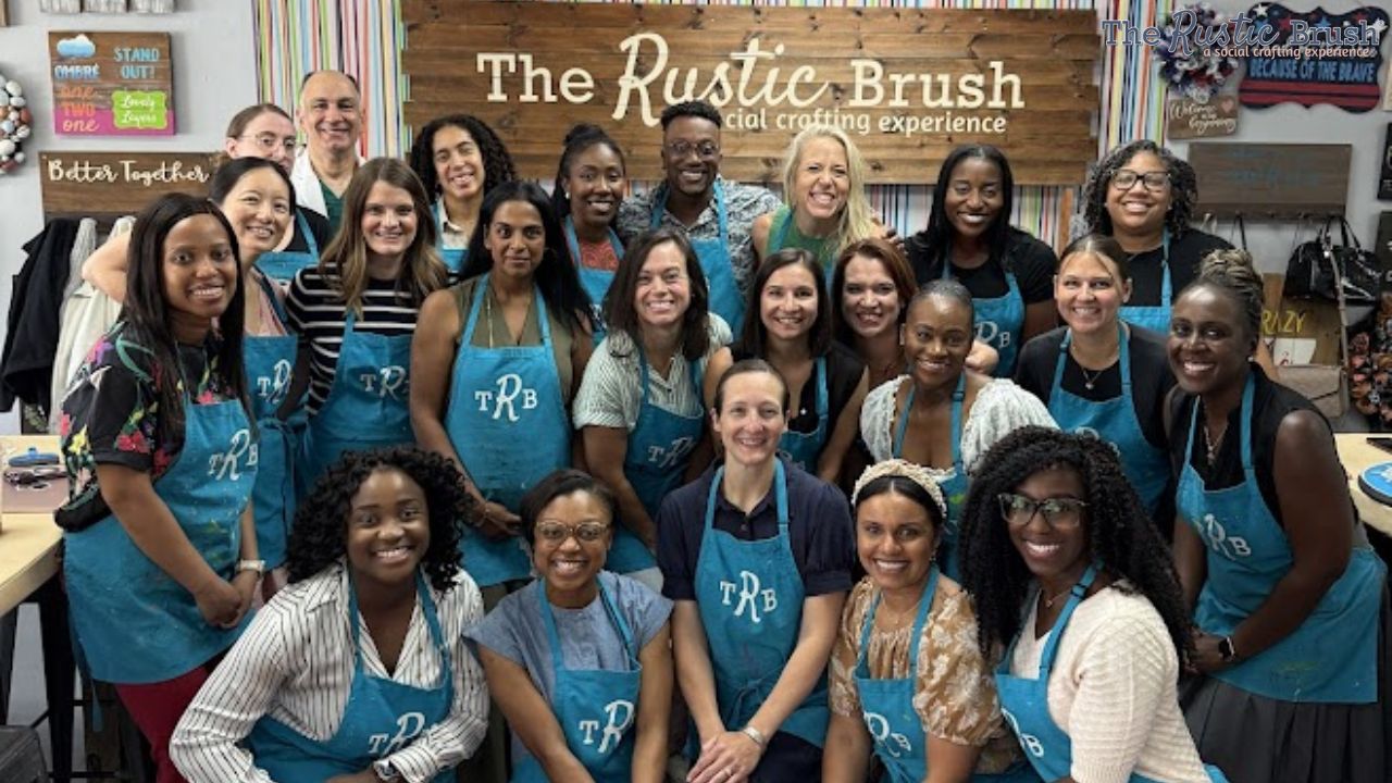 adult-beginner-art-classes Group of people smiling, wearing blue aprons in front of a wooden sign that reads 'The Rustic Brush'.