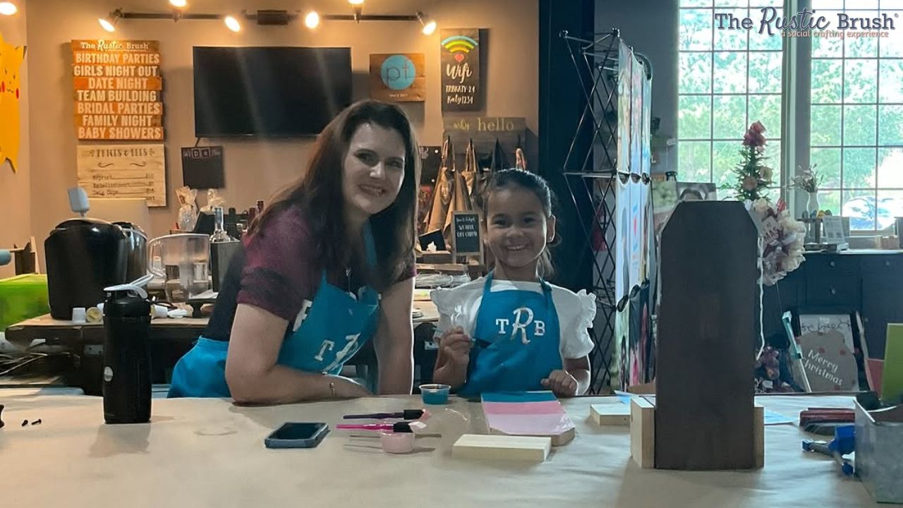 art-classes-for-adults-near-me (1) Two people in blue aprons smiling at a crafting workshop table.