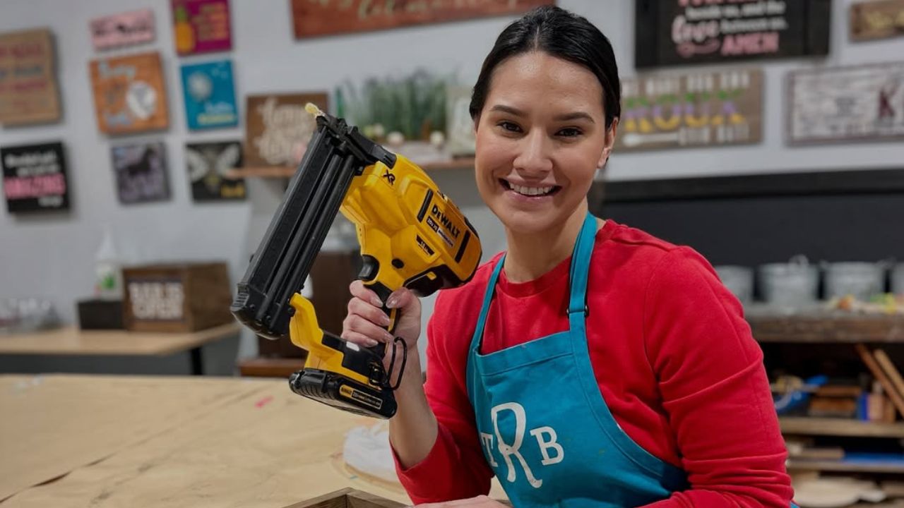 Smiling person in apron holding a yellow power tool in a decorated workshop.