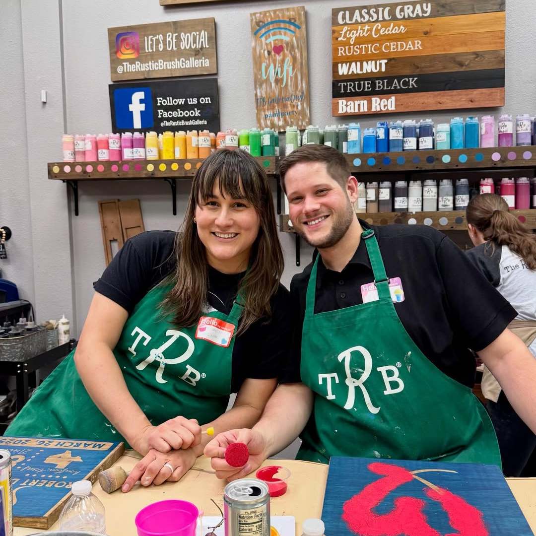 Two people wearing green aprons pose at a crafting table, surrounded by colorful paint bottles.