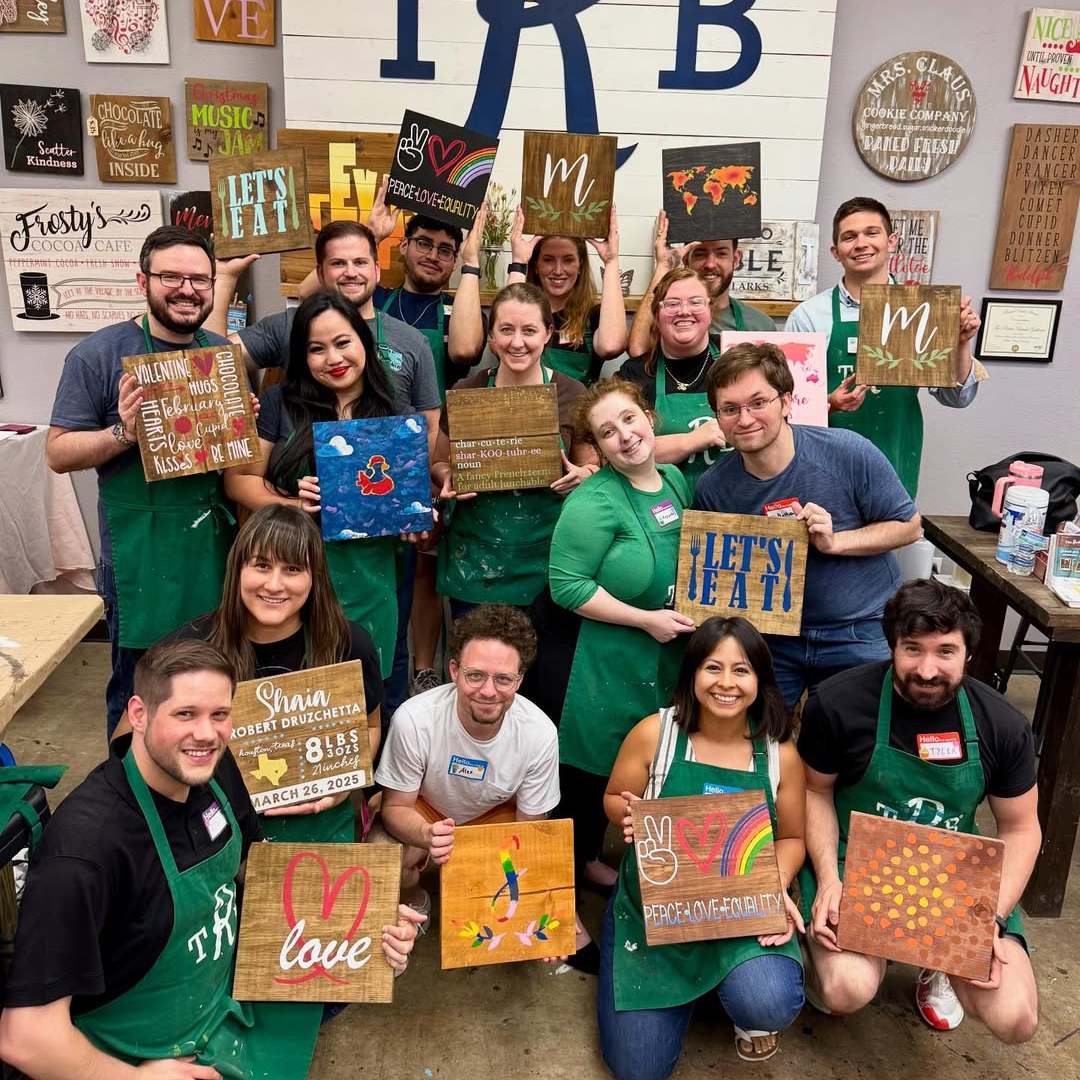 Group of people in green aprons holding handmade art boards, smiling in a workshop room.