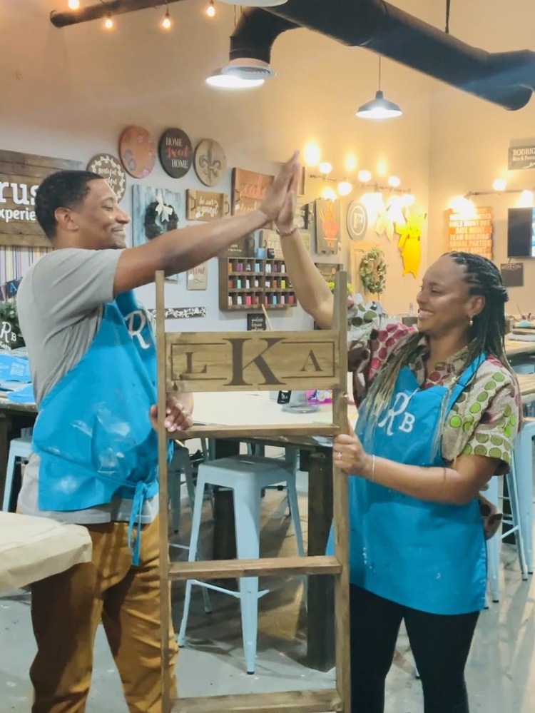 Two people in blue aprons high-five while holding a wooden frame indoors.