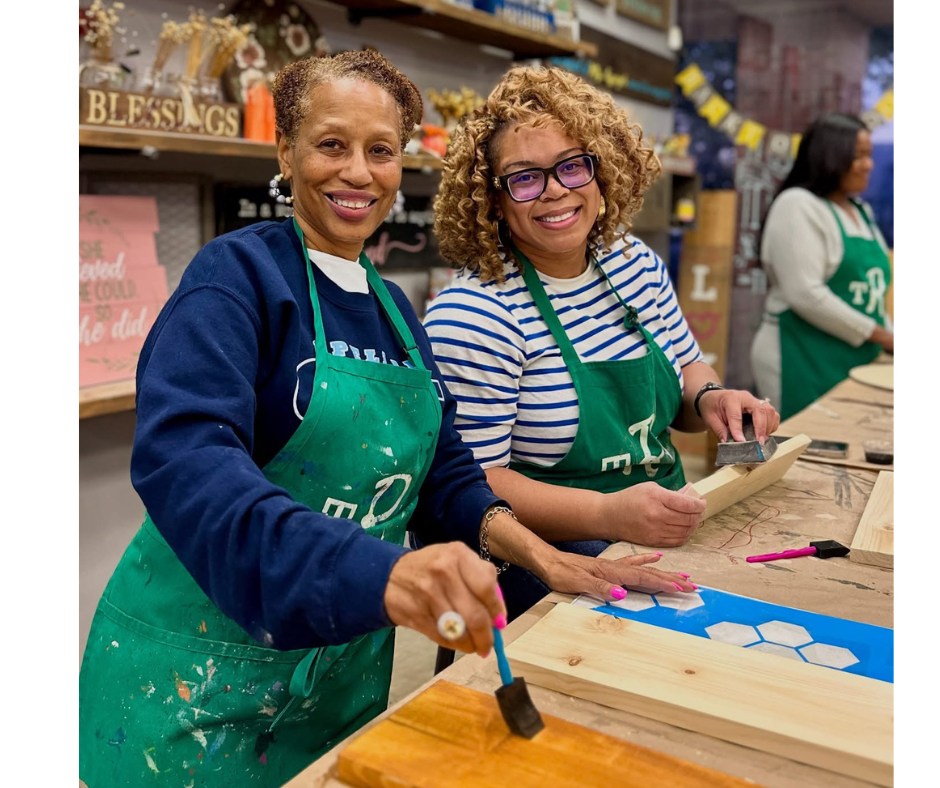 Two women crafting with paint in a workshop, wearing green aprons and smiling.