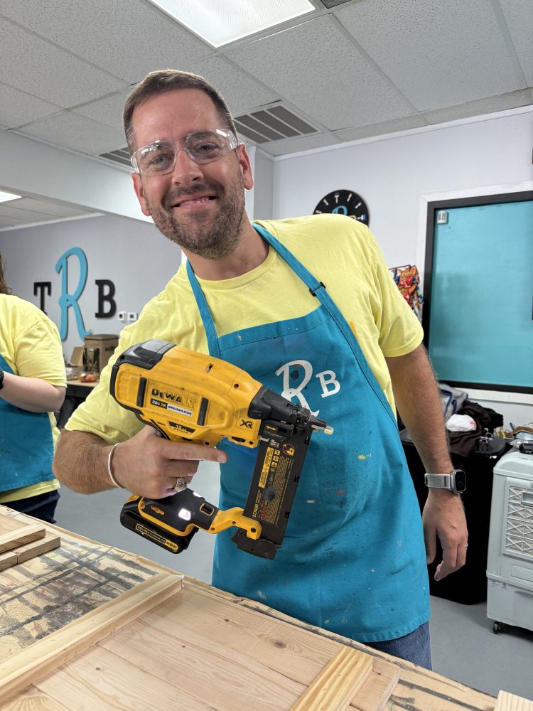 Man in safety glasses and apron holding a power tool in a workshop.