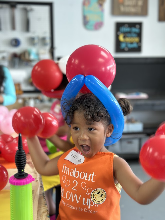 Child with balloon hat holding red balloons, wearing orange apron with text.