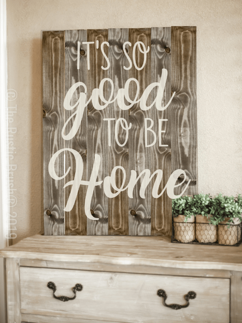 Wooden sign with 'It's so good to be home' beside small potted plants on a dresser.