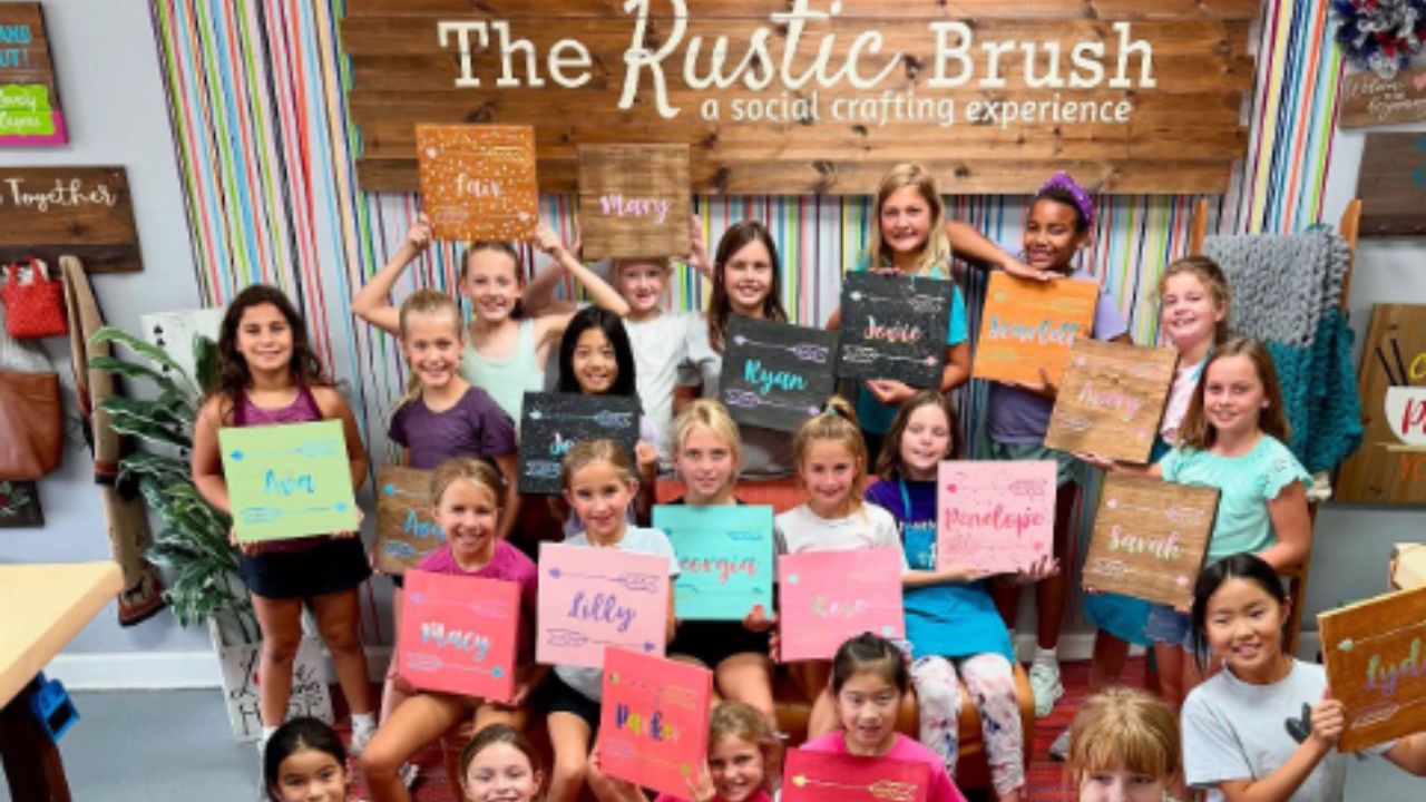 Group of kids holding colorful signs at a craft studio, 'The Rustic Brush' visible in the background.