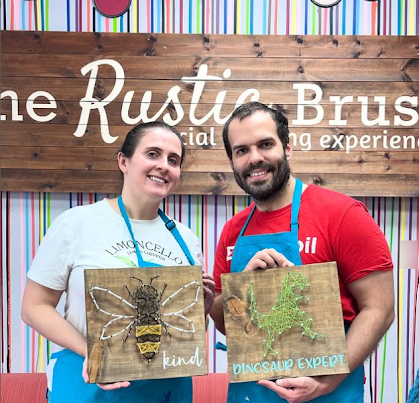 Two smiling people holding crafted wooden boards with bee and dinosaur designs in a creative studio.