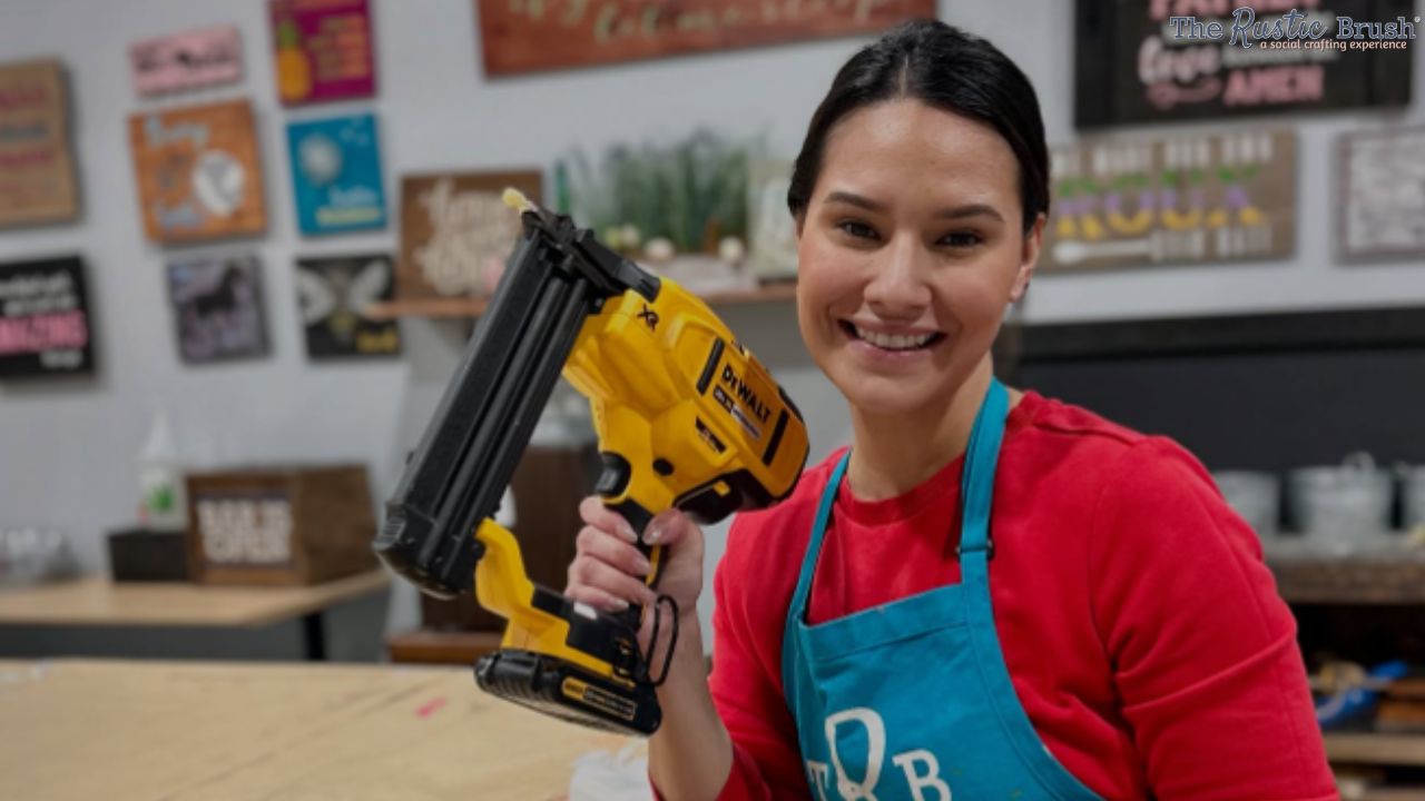 Woman smiling, holding a yellow nail gun, in a craft studio.