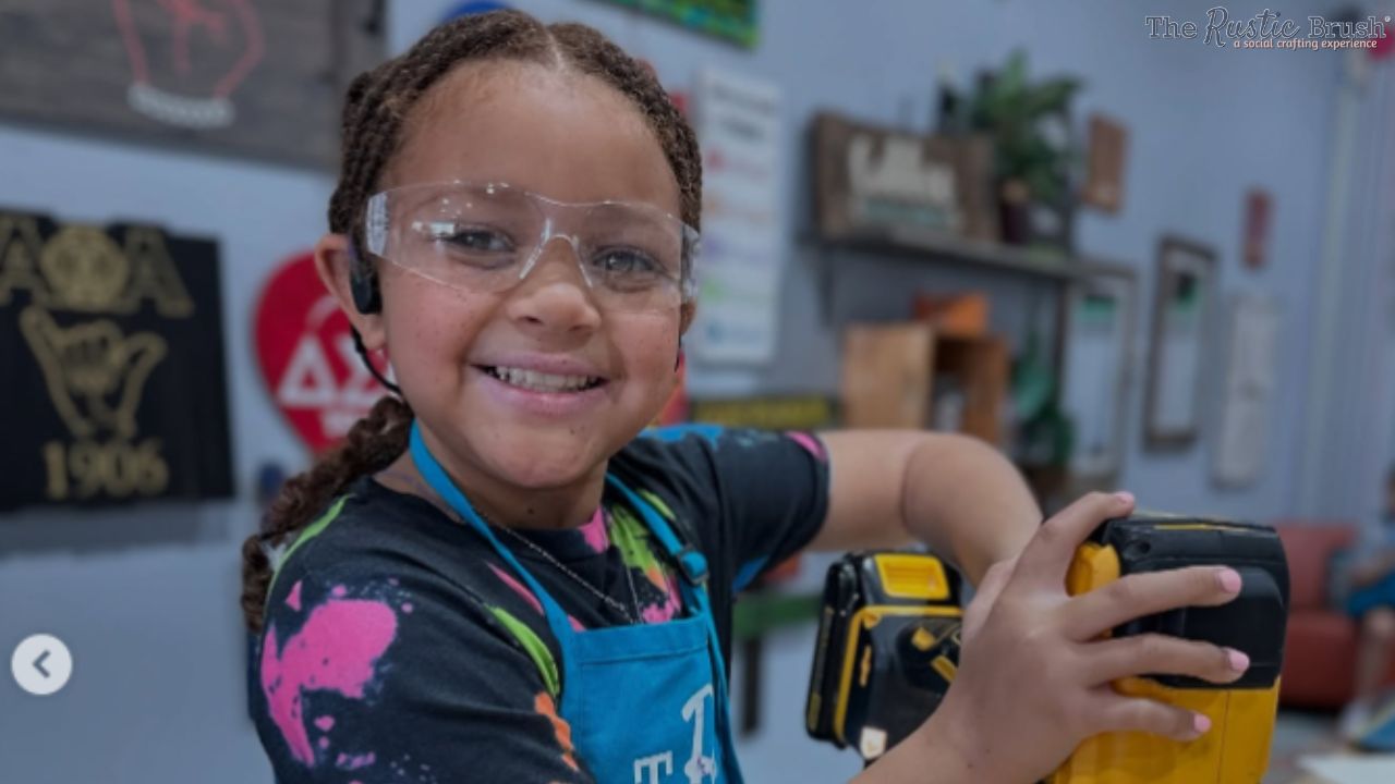 Child wearing goggles holding a yellow power drill in a workshop setting.