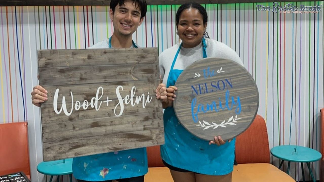 Two people smiling, holding wooden signs with painted text in art studio.