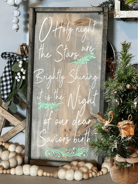 Wooden sign with 'O Holy Night' lyrics, greenery accents beside a small Christmas tree.