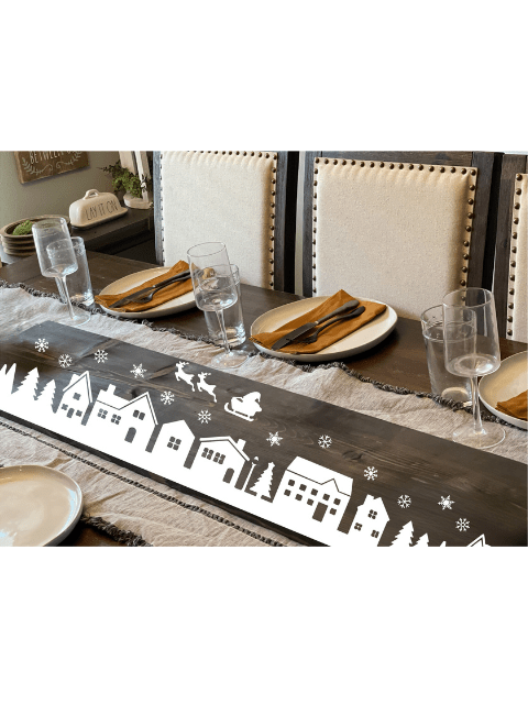 Dining table set with plates, glasses, and a runner decorated with a winter village scene.