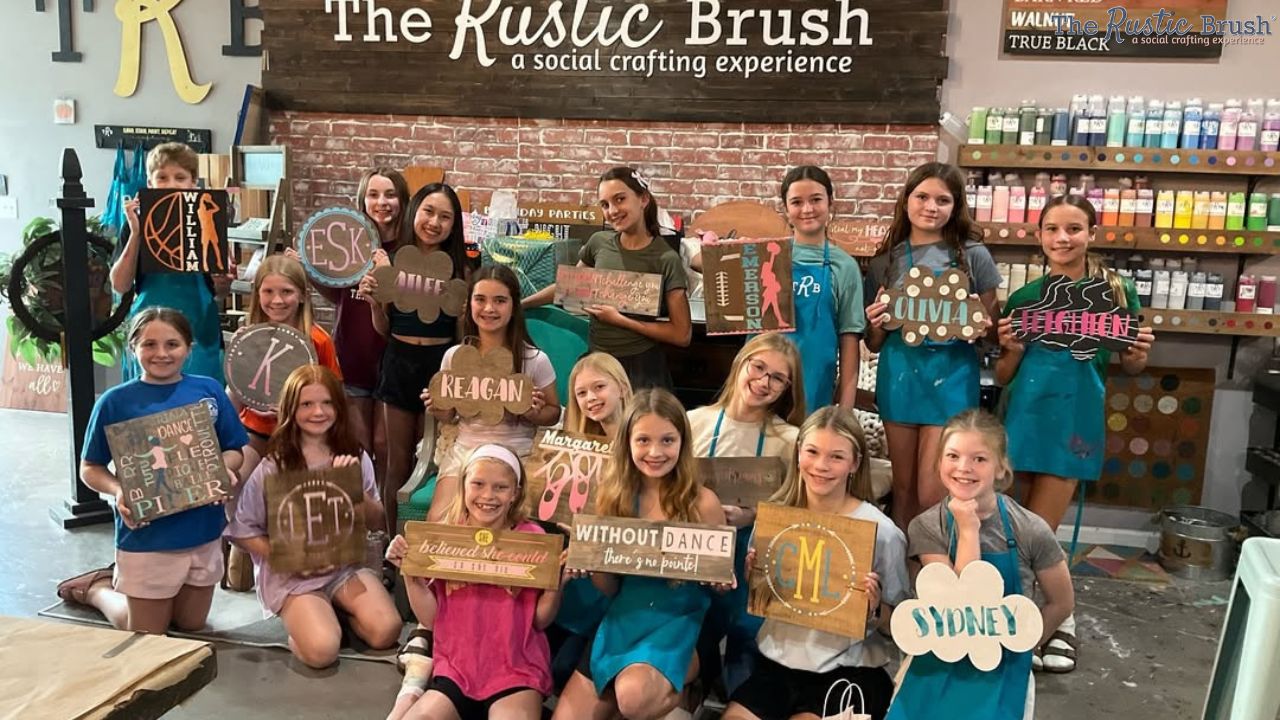 Group of children holding crafts in a workshop with a sign reading 'The Rustic Brush' in the background.