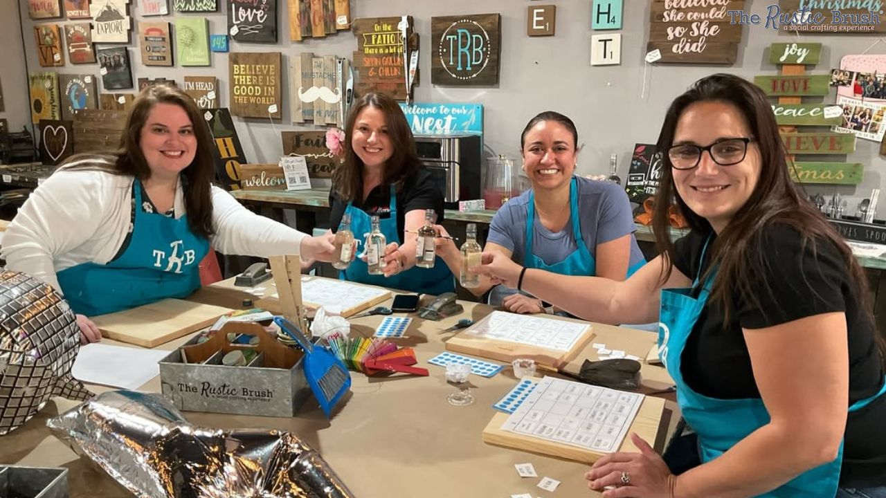 Four women smiling and toasting with drinks at a craft workshop, surrounded by wooden signs.