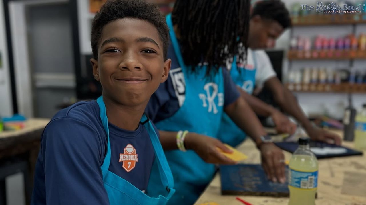 Smiling boy in blue apron, with others crafting at a workshop.