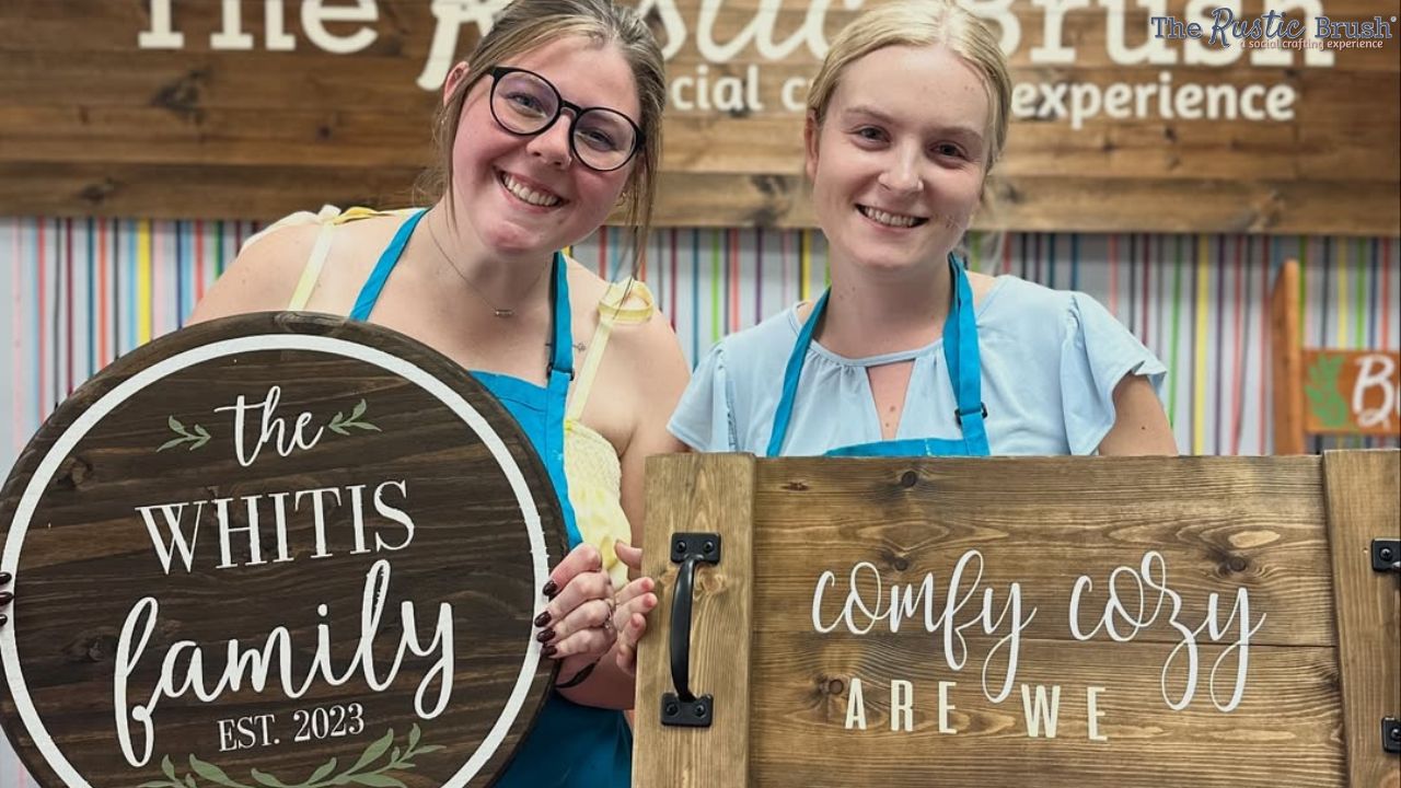 Two women smiling, holding decorative wooden signs in a craft studio.