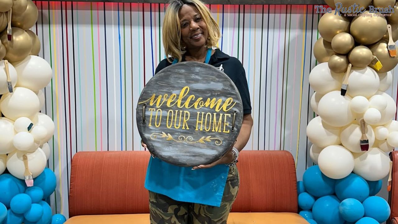 Person holding a round wooden sign saying 'Welcome to Our Home' with balloon decorations around.