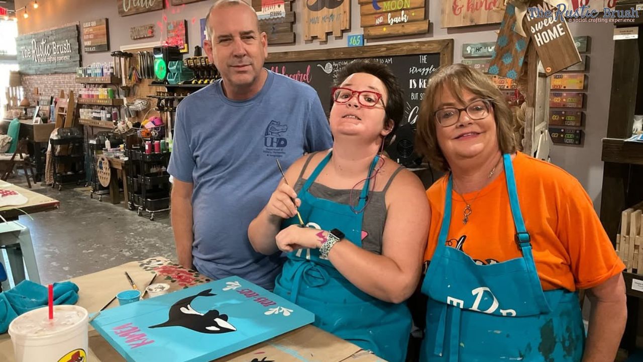 Three people in an art studio, two wearing aprons, one holding a paintbrush, surrounded by craft supplies.