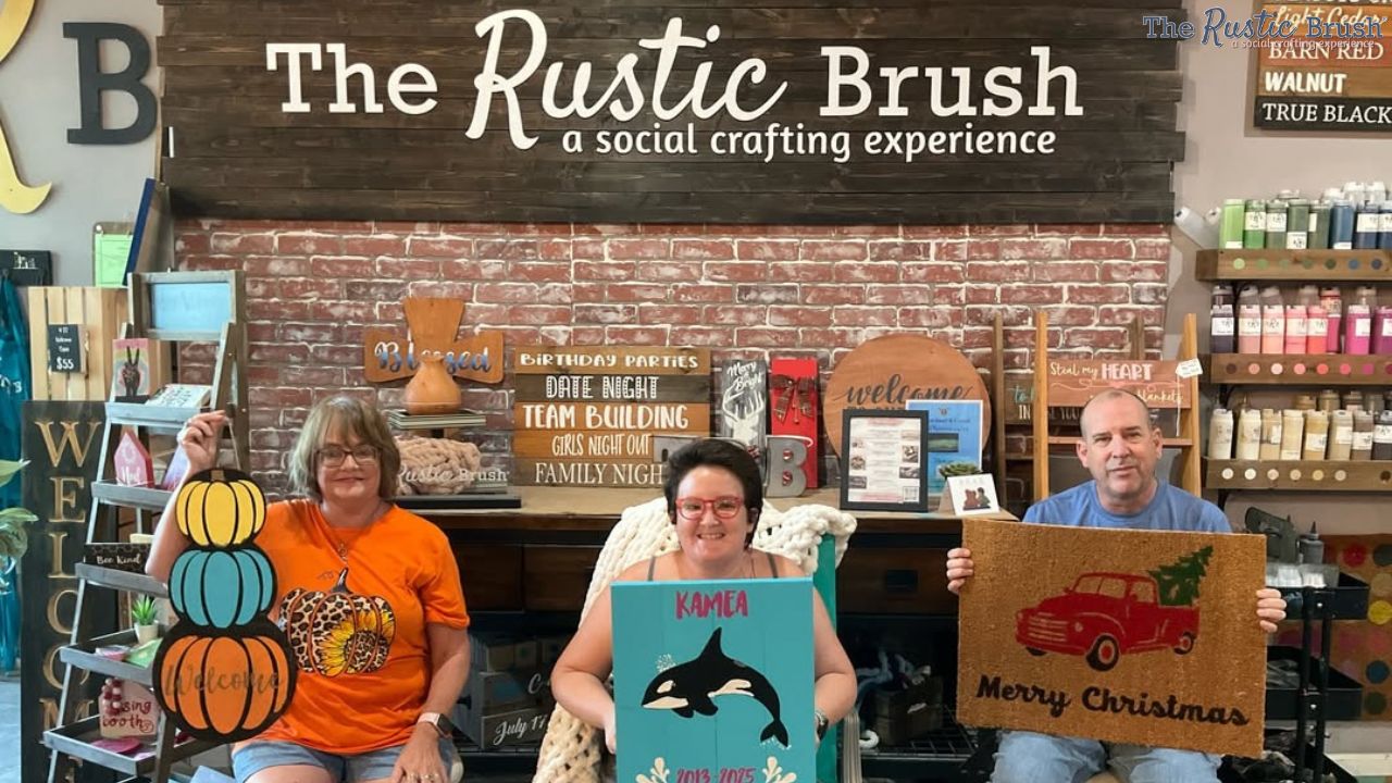 Three people holding crafts at The Rustic Brush store with decorative wood and paint supplies in the background.