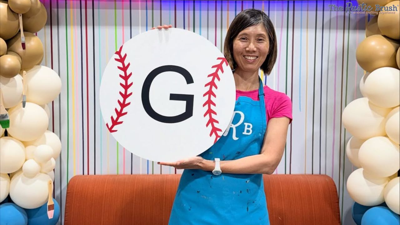 Woman holds a large sign resembling a baseball with a 'G' on it, standing in front of balloon decorations.