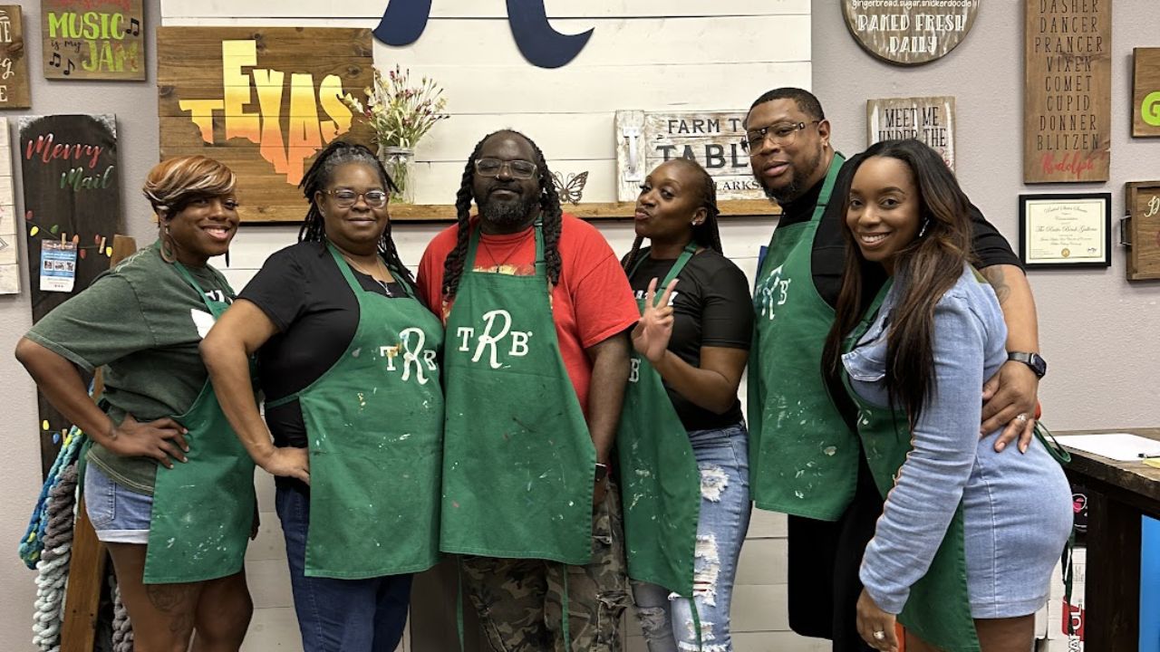 A group of adults inside a crafting studio, posing beneath a large “TRB” wall sign, with handmade wooden décor displayed around them.