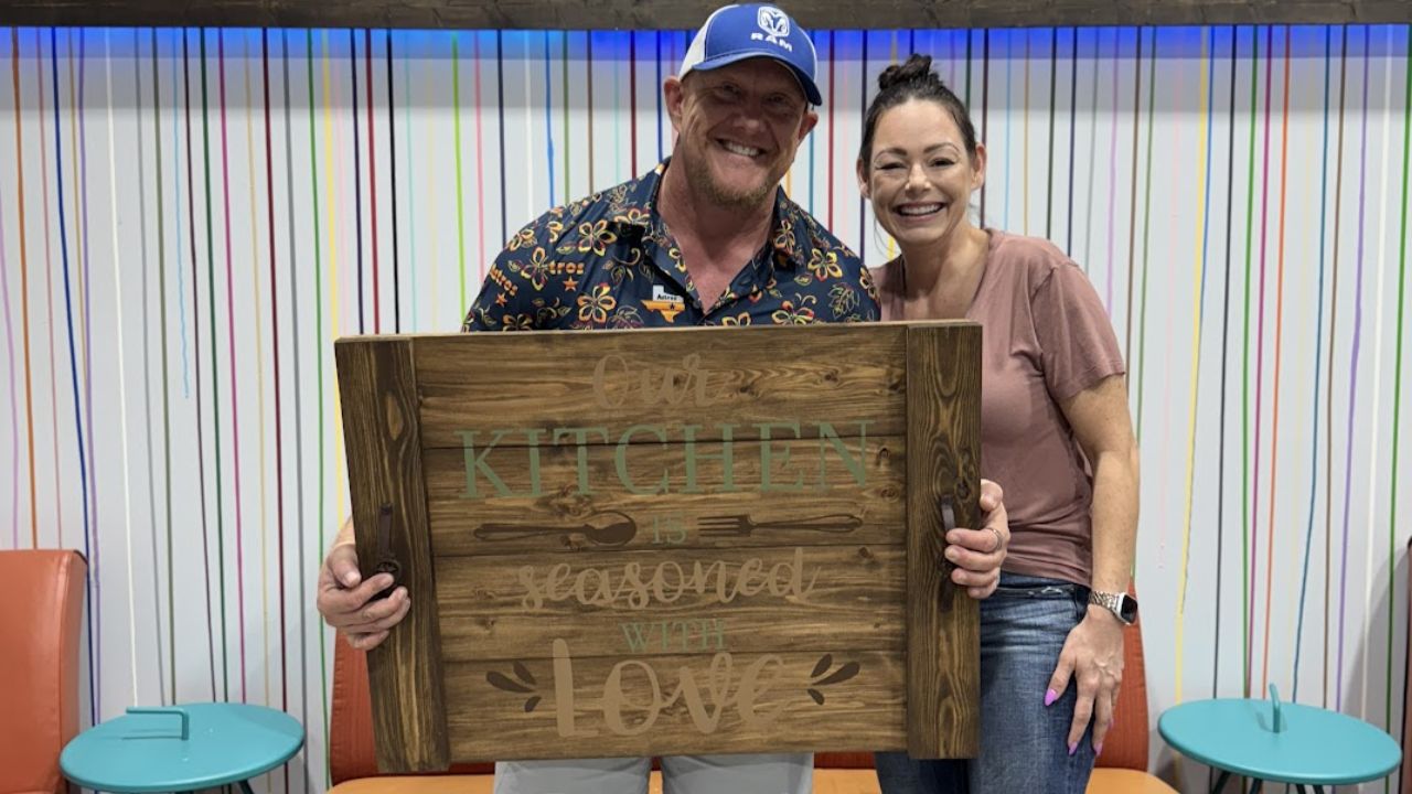 Smiling man and woman holding a wooden kitchen sign with colorful striped background.