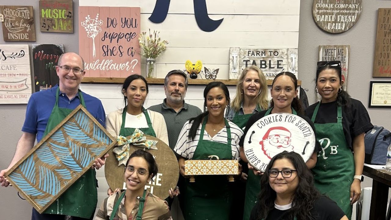 Nine people in aprons holding decorative signs, smiling, in front of wall art.