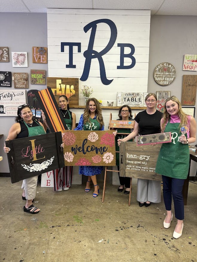 Six people smiling and holding handmade signs in front of a decorated wall with 'TRB' letters.