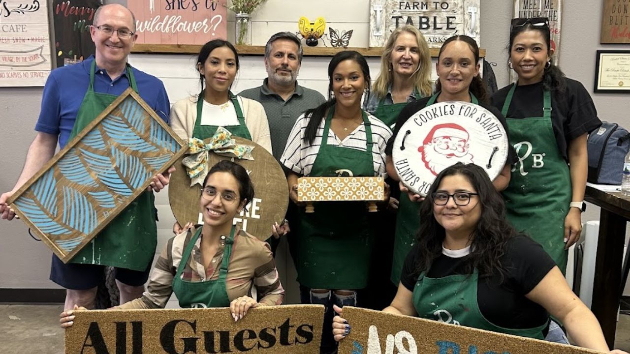 Group of people wearing aprons holding decorative signs and craft items in a workshop setting.